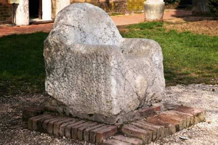 Sedia monumentale in pietra antica, decorativa e storica, posizionata su un basamento di mattoni, situata in un incantevole giardino con erba verde e piante.