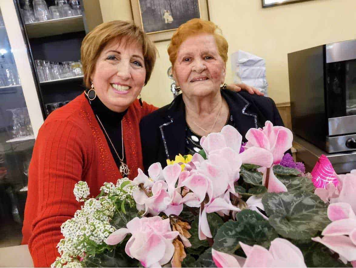 Fiori di ciclamino rosa in primo piano con due donne anziane sorridenti sullo sfondo, ambientazione cucina o ristorante.