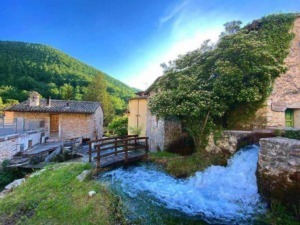Prezzo di una pittoresca scena di un villaggio di montagna italiano con case di pietra e un torrente che scorre, immersa nella natura e nel verde, ideale per blog di viaggi e turismo rurale.