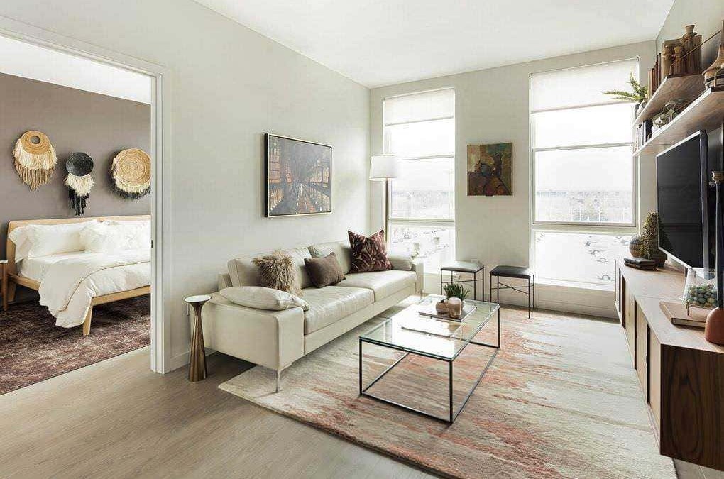 Luminoso soggiorno con grandi finestre, divano beige con cuscini decorativi, tavolino in vetro e scaffale con decorazioni in stile moderno, ideale per ambienti di casa accoglienti e di tendenza.
