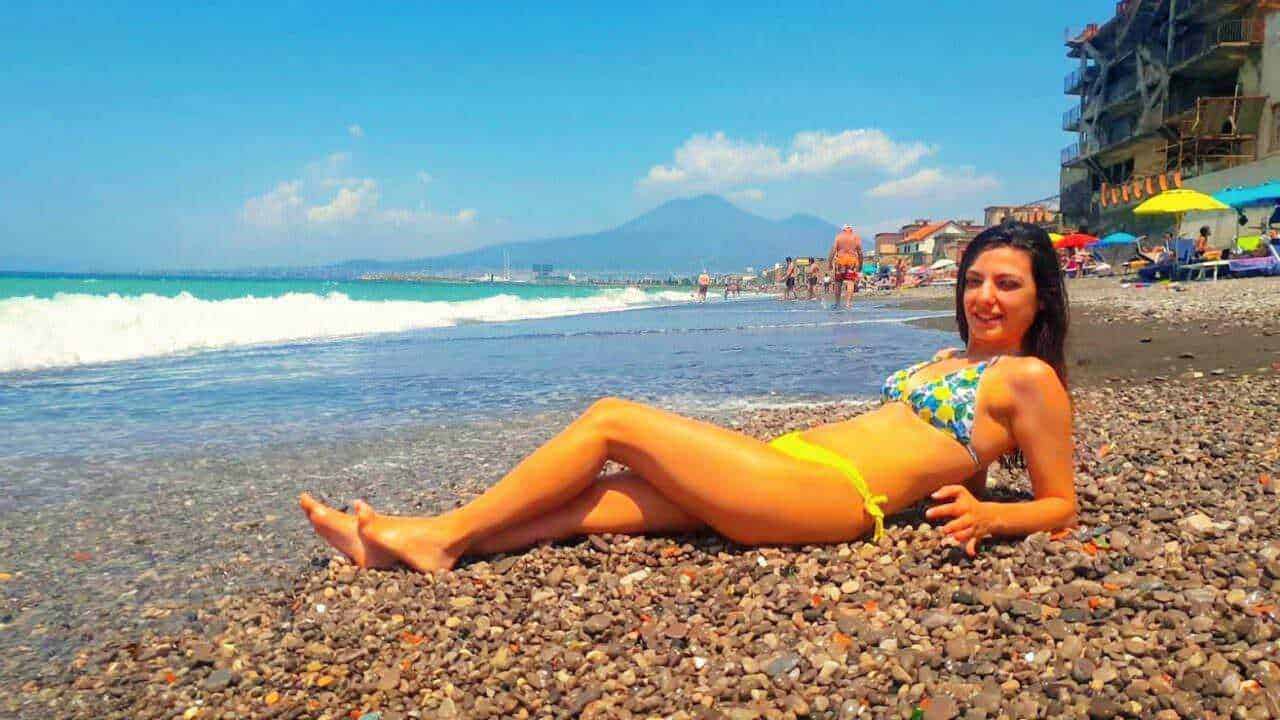 Spiaggia al mare con donna in bikini sdraiata sui ciottoli, ombrelloni colorati e cielo soleggiato, vista sul mare e sulle montagne sullo sfondo.