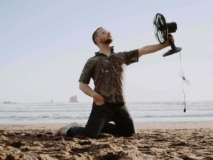 Fan da spiaggia con uomo che tiene in mano un ventilatore portatile, quindi foto di relax e comfort estivi, ambientazione naturale mare, resistenza al caldo