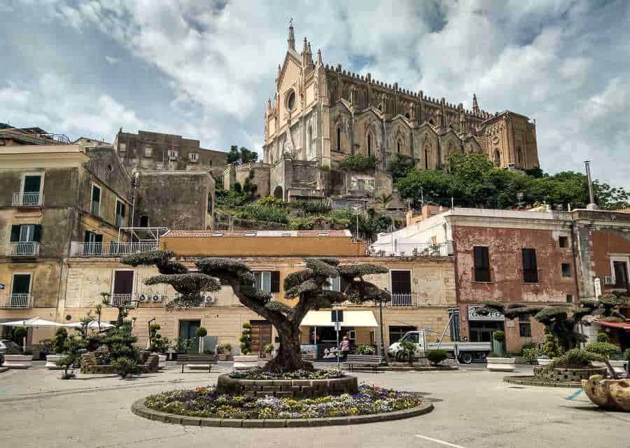 Fontana centrale con alberi sagomati e fiori in primo piano, sullo sfondo una chiesa storica su una collina, suggestiva scena urbana con edifici colorati e cielo nuvoloso, immagine di suggestiva piazza italiana con monumento e architettura storica.