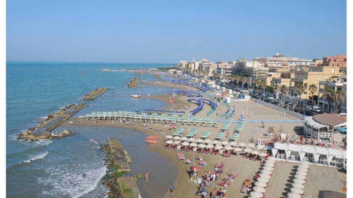 Spiaggia affollata con ombrelloni e persone al mare, città sul lungomare con bar e strutture ricettive, blu cielo sereno, ambiente estivo e turistico ai lidi.