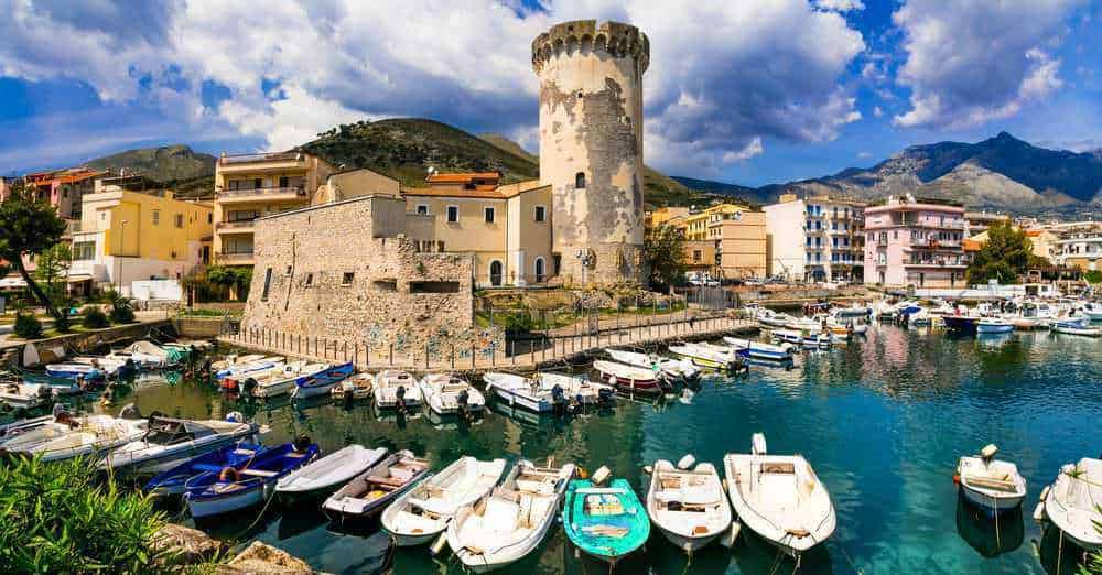 Castello sul porto di un paesino italiano, con barche e abitazioni colorate, scenario di turismo e bellezze costiere del Mediterraneo.