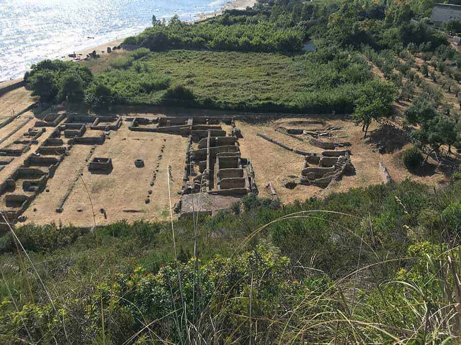 Antica area archeologica con resti di mura e strutture in pietra, vista dalla collina, circondata da vegetazione e vicino al mare, sito di interesse storico e culturale in Italia, esploriamo le rovine storiche.