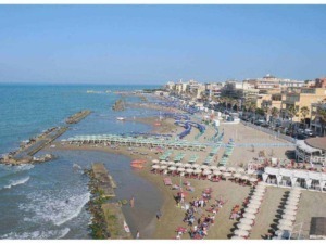 Spiaggia affollata con ombrelloni e persone al mare, città sul lungomare con bar e strutture ricettive, blu cielo sereno, ambiente estivo e turistico ai lidi.