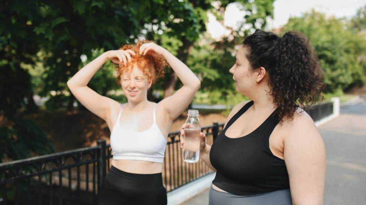 Passeggiata all'aperto tra due donne felici e sorridenti, una che ride e si sistema i capelli, l'altra con una bottiglia d'acqua.