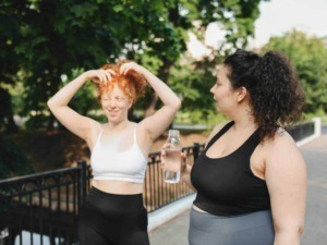 Passeggiata all'aperto tra due donne felici e sorridenti, una che ride e si sistema i capelli, l'altra con una bottiglia d'acqua.