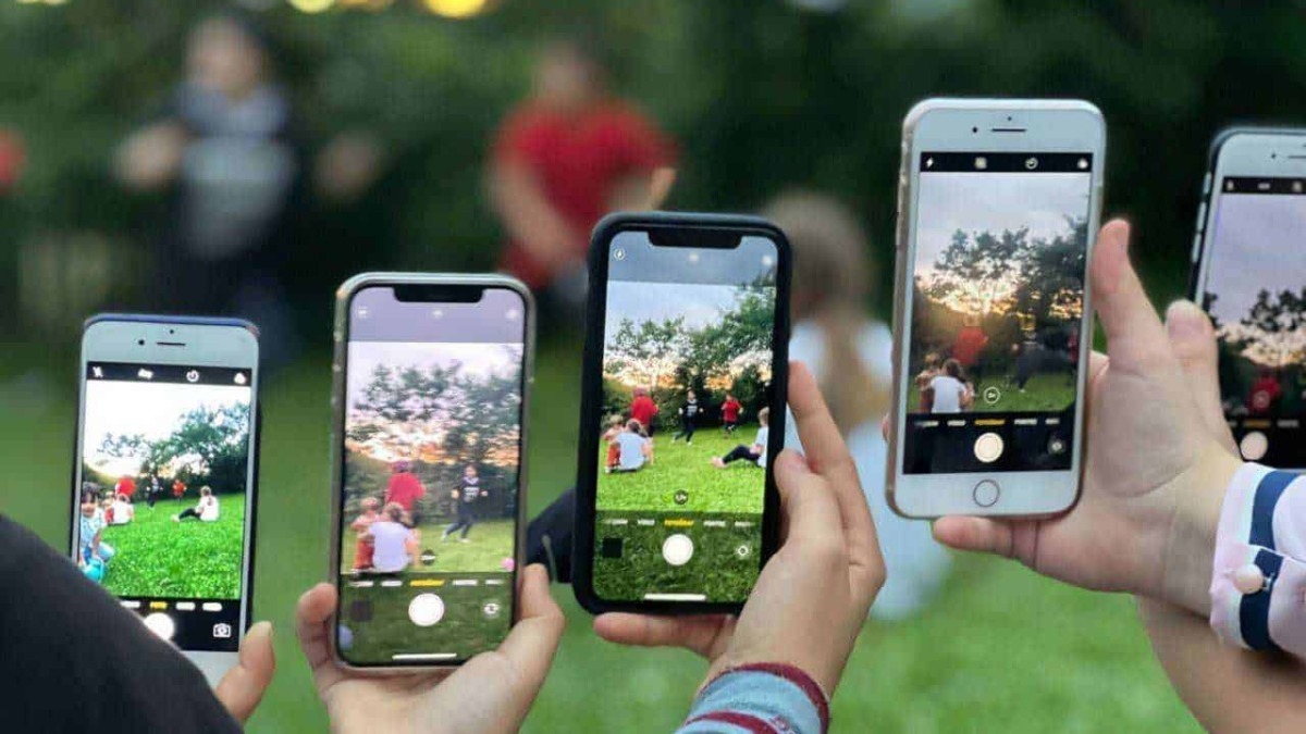 Digital camera capturing people taking photos with smartphones during outdoor event, sunset sky in background, greenery, and social gathering atmosphere.