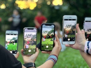 Digital camera capturing people taking photos with smartphones during outdoor event, sunset sky in background, greenery, and social gathering atmosphere.