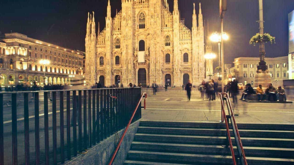 Cattedrale di Milano illuminata di notte con persone che passeggiano e seduti sulla piazza. Scenario urbano affascinante con luci calde, architettura gotica e atmosfera vivace e tranquilla.