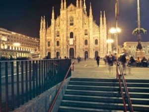 Cattedrale di Milano illuminata di notte con persone che passeggiano e seduti sulla piazza. Scenario urbano affascinante con luci calde, architettura gotica e atmosfera vivace e tranquilla.