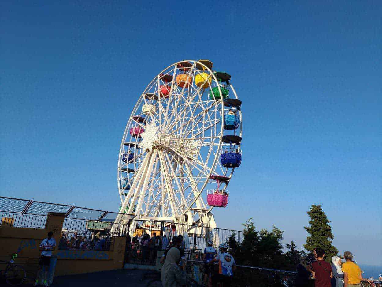 Ruota panoramica colorata in un parco di divertimenti all'aperto con visitatori che la ammirano sotto un cielo sereno, perfetta per articoli su attrazioni turistiche e divertimento.