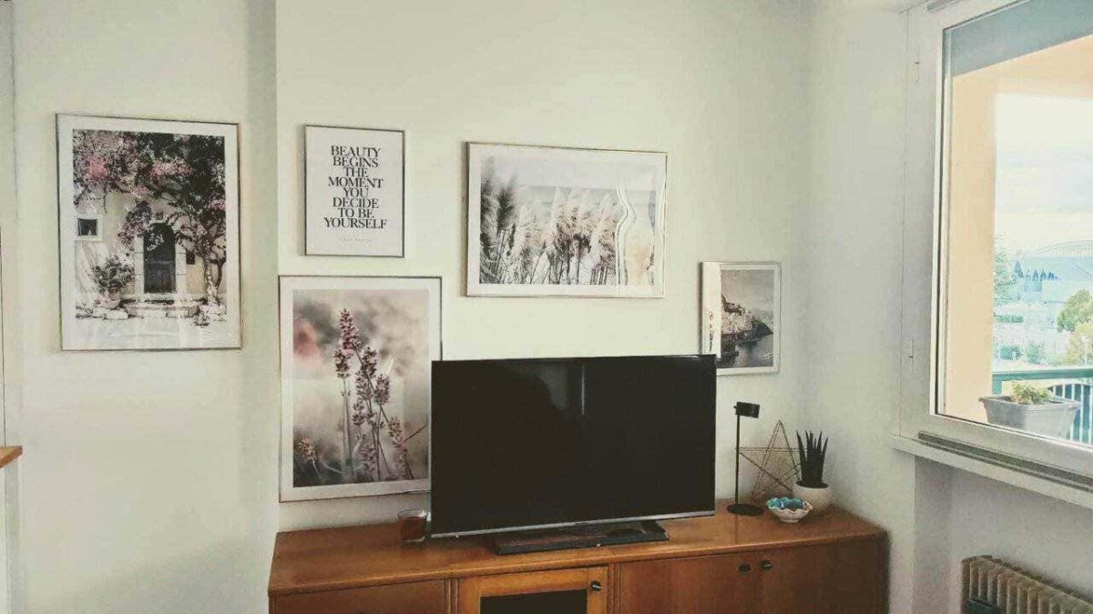 Fotografia di un soggiorno con parete decorata da quadri astratti, stampe di fiori e citazioni motivazionali, arredato con mobile in legno, televisore, pianta e oggetti decorativi.