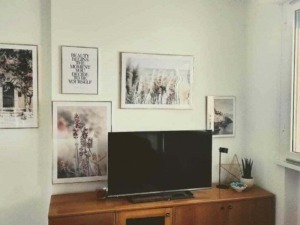 Fotografia di un soggiorno con parete decorata da quadri astratti, stampe di fiori e citazioni motivazionali, arredato con mobile in legno, televisore, pianta e oggetti decorativi.