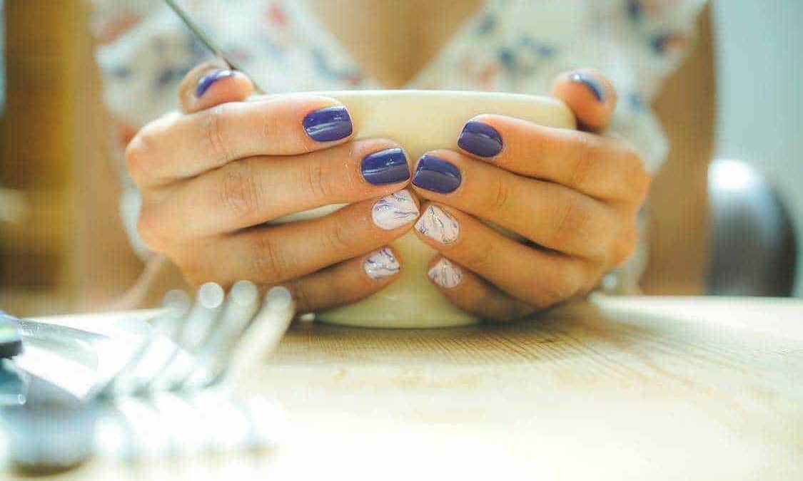 Manicure elegante con unghie colorate di viola e decorazioni artistiche, mani che tengono una tazza di tè o caffè in un'atmosfera rilassante e raffinata, perfetta per blog di bellezza e stile di vita.