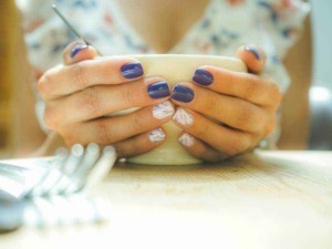 Manicure elegante con unghie colorate di viola e decorazioni artistiche, mani che tengono una tazza di tè o caffè in un'atmosfera rilassante e raffinata, perfetta per blog di bellezza e stile di vita.