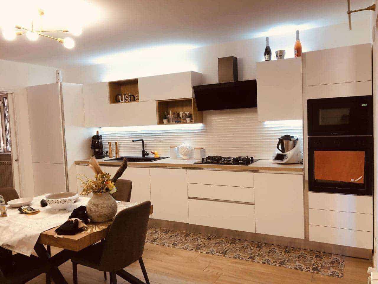 Modern cucina bianca con elettrodomestici integrati, piano cottura a gas e area pranzo con tavolo decorato con centro tavola floreale e sedie imbottite. Atmosfera luminosa e accogliente.