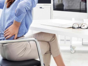 Dolore alla schiena durante il lavoro in ufficio, donna seduta con mano sulla schiena, problemi di postura, mal di schiena da ufficio, ergonomia sul lavoro, salute e benessere al lavoro.