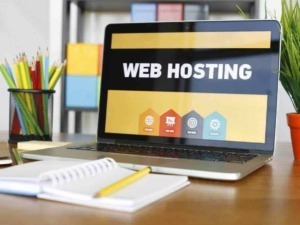 Immagine di un laptop con schermo che mostra "Web Hosting" in un ambiente di lavoro moderno e colorato, con colori vivaci e oggetti di cancelleria intorno.