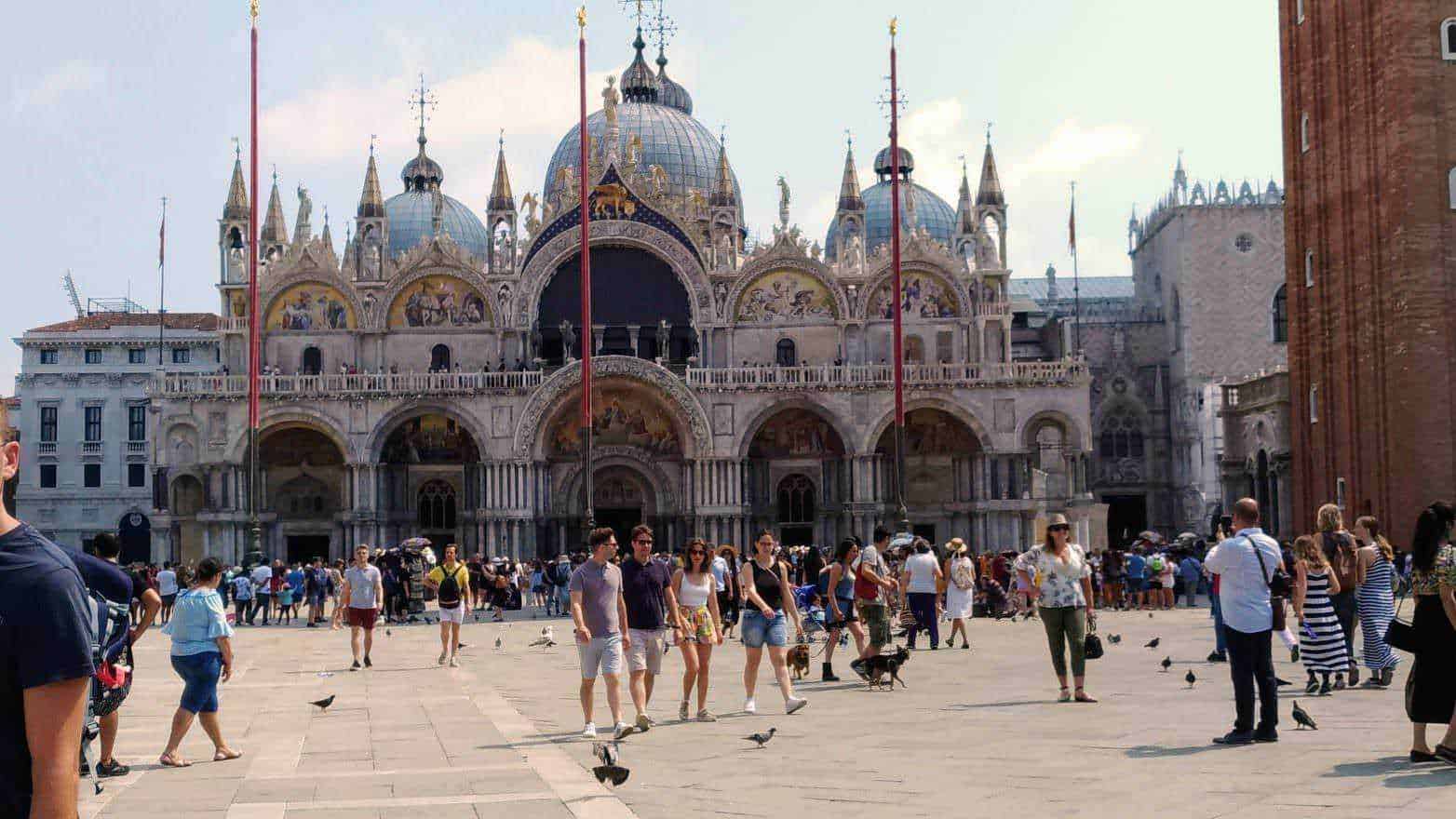 Weekend a Venezia: 13 cose che non potete perdervi! 2 Altezza della basilica di San Marco a Venezia, piazza affollata con turisti e piccioni, vista dettagliata dell'architettura storica e decorazioni artistiche del monumento.