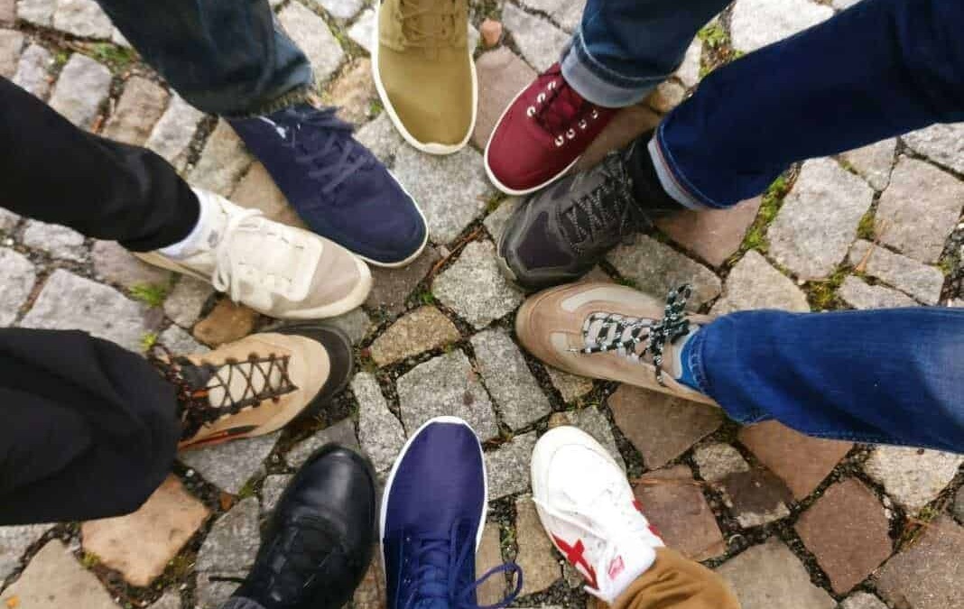 Scarpe sportive di diversi stili e colori disposte a cerchio su un pavimento in pietra. Diversi amici si incontrano per condividere un momento di convivialità.