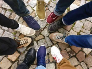 Scarpe sportive di diversi stili e colori disposte a cerchio su un pavimento in pietra. Diversi amici si incontrano per condividere un momento di convivialità.