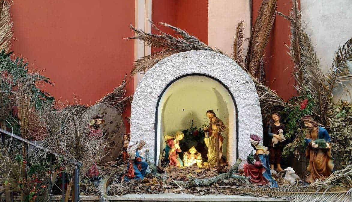 Presepe natalizio con statue di Gesù, Maria, Giuseppe e altri personaggi, decorato con piante secche e rami, posizionato in un ambiente esterno con sfondo di pareti colorate.