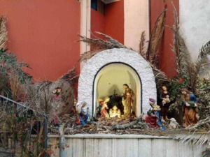 Presepe natalizio con statue di Gesù, Maria, Giuseppe e altri personaggi, decorato con piante secche e rami, posizionato in un ambiente esterno con sfondo di pareti colorate.