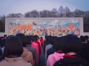 Particolare di una grande murale colorata rappresentante una manifestazione celebrativa, con molte persone che sventolano bandiere e ascoltano un discorso di una figura politica sullo sfondo naturale.