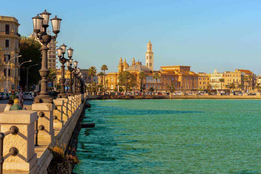 Panoramica serale del lungomare di Bari con palazzi storici illuminati e barche sul mare.