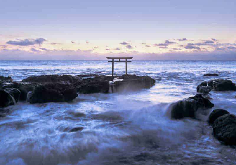 Torre shinto su scogliata marina durante tramonto, simbolo culturale giapponese con paesaggio marino in lontananza.
