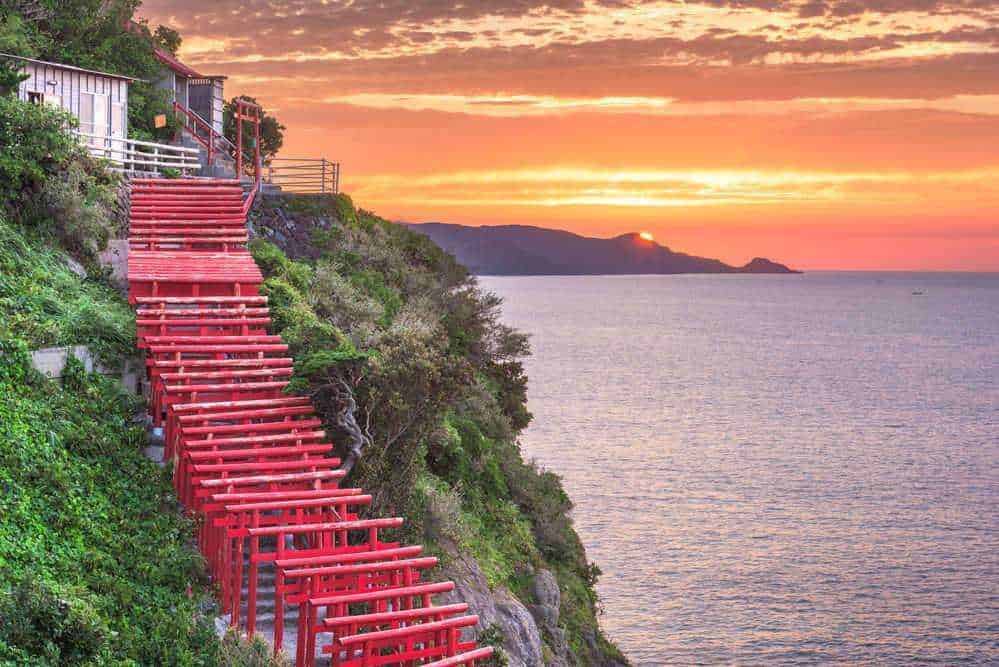 Scale di scale in legno rosso sulla scogliera con vista mare al tramonto, paesaggio costiero giapponese, atmosfera serena, natura e cultura, immagini di viaggi e viaggio, natura e paesaggi, bellezza naturale dell'oceano, attrazioni turistiche, escursioni e trekking, fotografie di paesaggi e natura, emozioni di viaggio e avventura.