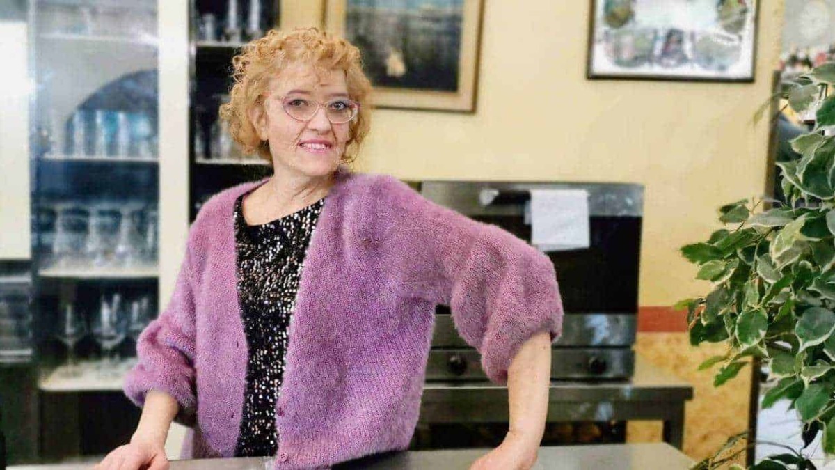 Vestito rosa, donna anziana con riccioli rossi e occhiali, sorridente, ambiente interno di un caffè o ristorante con piante e quadri alle pareti, lifestyle e social media.