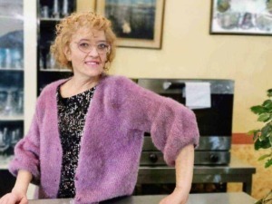 Vestito rosa, donna anziana con riccioli rossi e occhiali, sorridente, ambiente interno di un caffè o ristorante con piante e quadri alle pareti, lifestyle e social media.