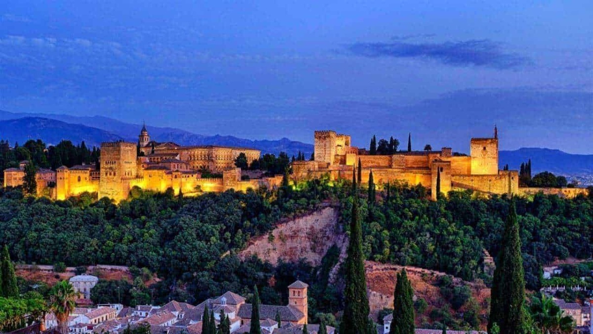 Castello di Alhambra illuminate di sera, vista panoramica della fortezza storica e delle mura, con paesaggi montani sullo sfondo, in Andalusia, Spagna.