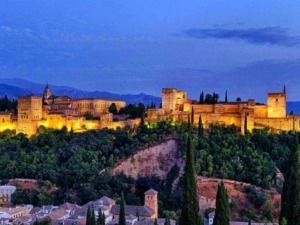 Castello di Alhambra illuminate di sera, vista panoramica della fortezza storica e delle mura, con paesaggi montani sullo sfondo, in Andalusia, Spagna.