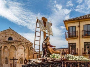 Festa religiosa con statua della Vergine Maria e Gesù morto, scenografiche decorazioni floreali e sfondo di edifici storici in pietra, cielo azzurro con nuvole.