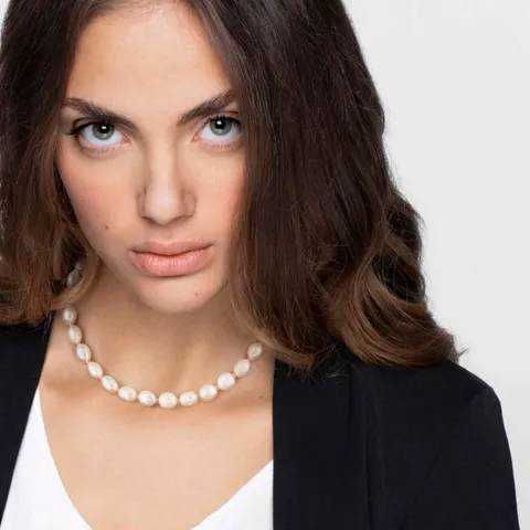 Ragazza con sguardo intenso, capelli castani e trucco naturale, indossa una collana di perle e un outfit elegante, perfetta per uno stile sofisticato e femminile.