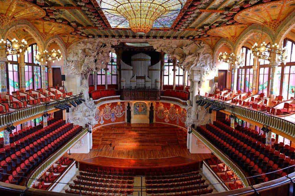 Modernismo catalano: Auditorium storico con palco in legno e decorazioni artistiche in stile Art Nouveau, illuminato da grandi vetrate colorate e lampadari eleganti. Perfetto per eventi culturali e concerti.