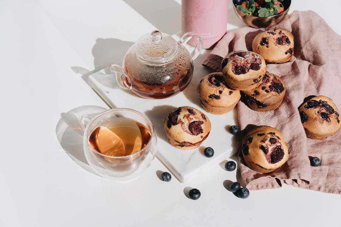 Dolci muffin ai mirtilli e tè fumante su tavola bianca con ciotola di frutta, perfetti per colazione o merenda, atmosfera luminosa e accogliente, ideale per ricette dolci fatte in casa.