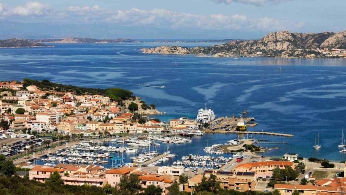 Vita marina e paesaggio costiero con porticciolo, imbarcazioni e case colorate, sotto cielo nuvoloso nella splendida isola mediterranea.