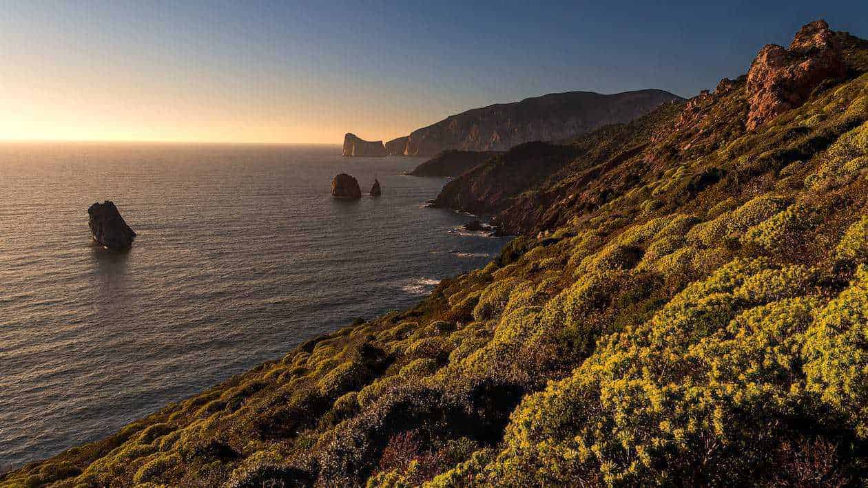 Itinerario Sardegna in Camper: dove andare? 3 Scogliere e coste del mare al tramonto con vegetazione mediterranea e spruzzi d'acqua, paesaggio naturale suggestivo e scenografico, destinazione ideale per escursioni e relax.