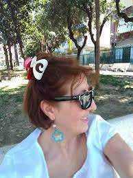 Immagine di una donna che si gode il sole in un parco con occhiali da sole e accessori colorati, ambientazione estiva con alberi e natura sullo sfondo.