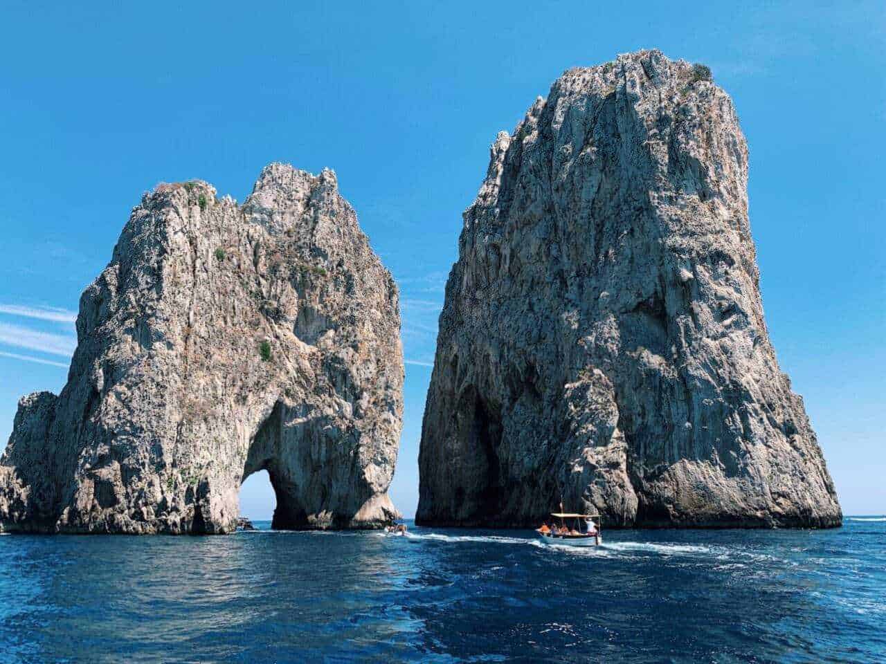 Maestose scogliere di pietra con arco naturale nel mare blu con vista sulla costa campana e barca che naviga nelle acque cristalline e cielo sereno e luminoso, ideale per promuovere viaggi go turismo in Italia.