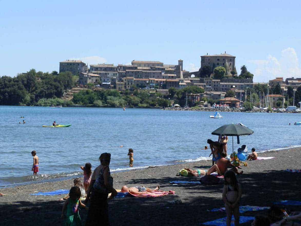 Spiaggia affollata con bambini e famiglie che si rilassano al sole, nuotano e praticano sport acquatici vicino a un castello storico sullo sfondo, tra mare e natura.