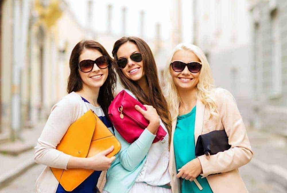 Vivian o ragazze alla moda con occhiali da sole sorridenti passeggiando in città, indossando abbigliamento trendy e accessori colorati, in un contesto urbano di giorno.