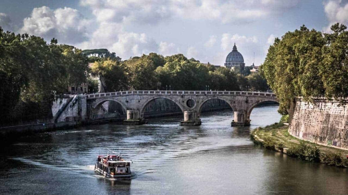 Pontes storiche sul Tevere a Roma con vista sul Vaticano e cielo nuvoloso. Nave che naviga sul fiume, circondato da alberi e edifici storici.