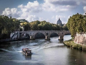 Pontes storiche sul Tevere a Roma con vista sul Vaticano e cielo nuvoloso. Nave che naviga sul fiume, circondato da alberi e edifici storici.